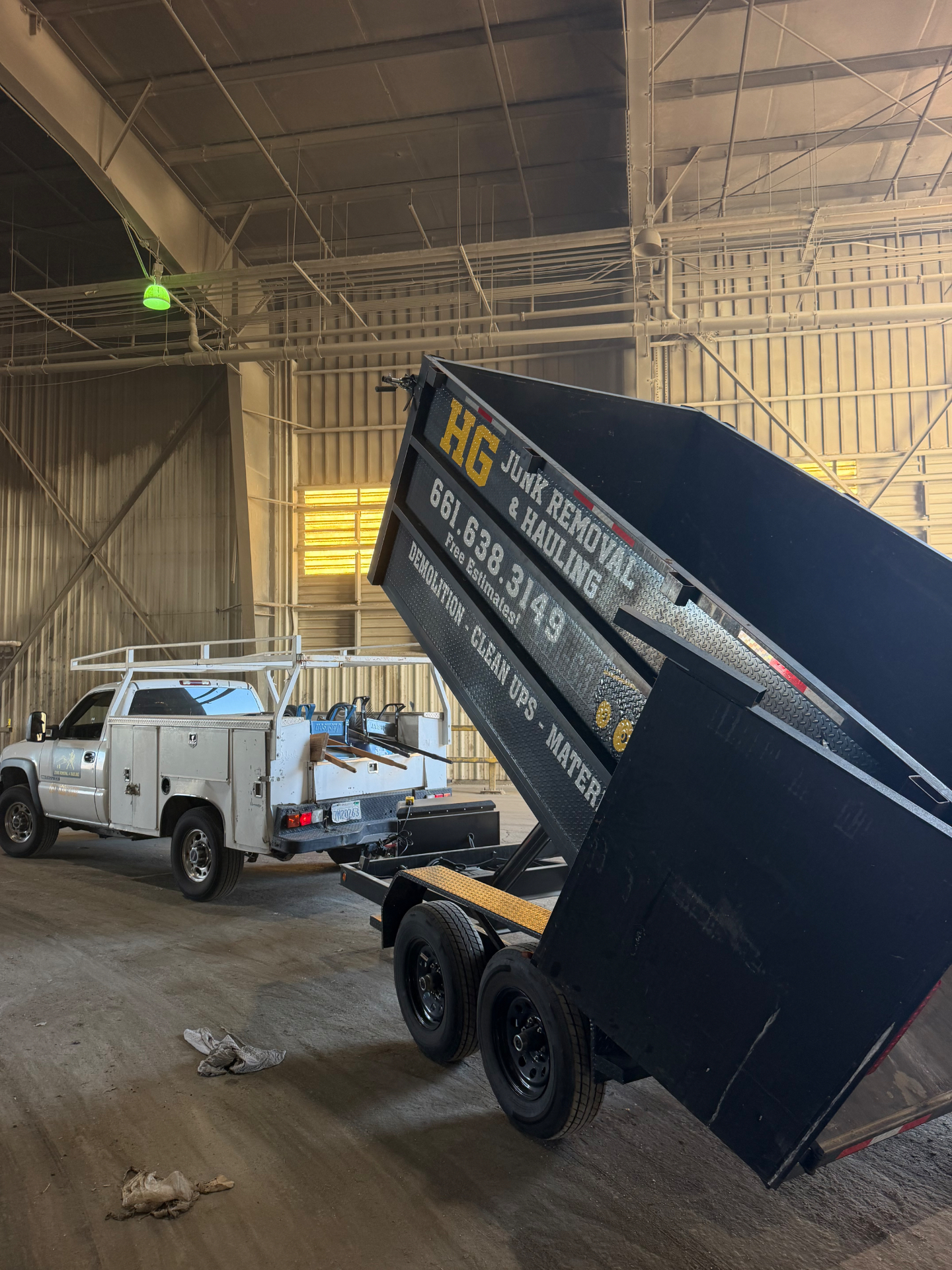 HG Junk Removal raised dump trailer and white work truck inside a commercial facility